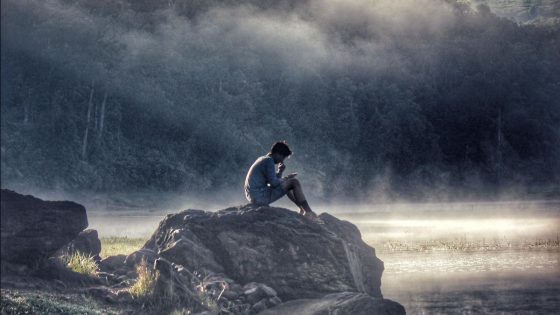photo of man sitting on rocks by water in the forest