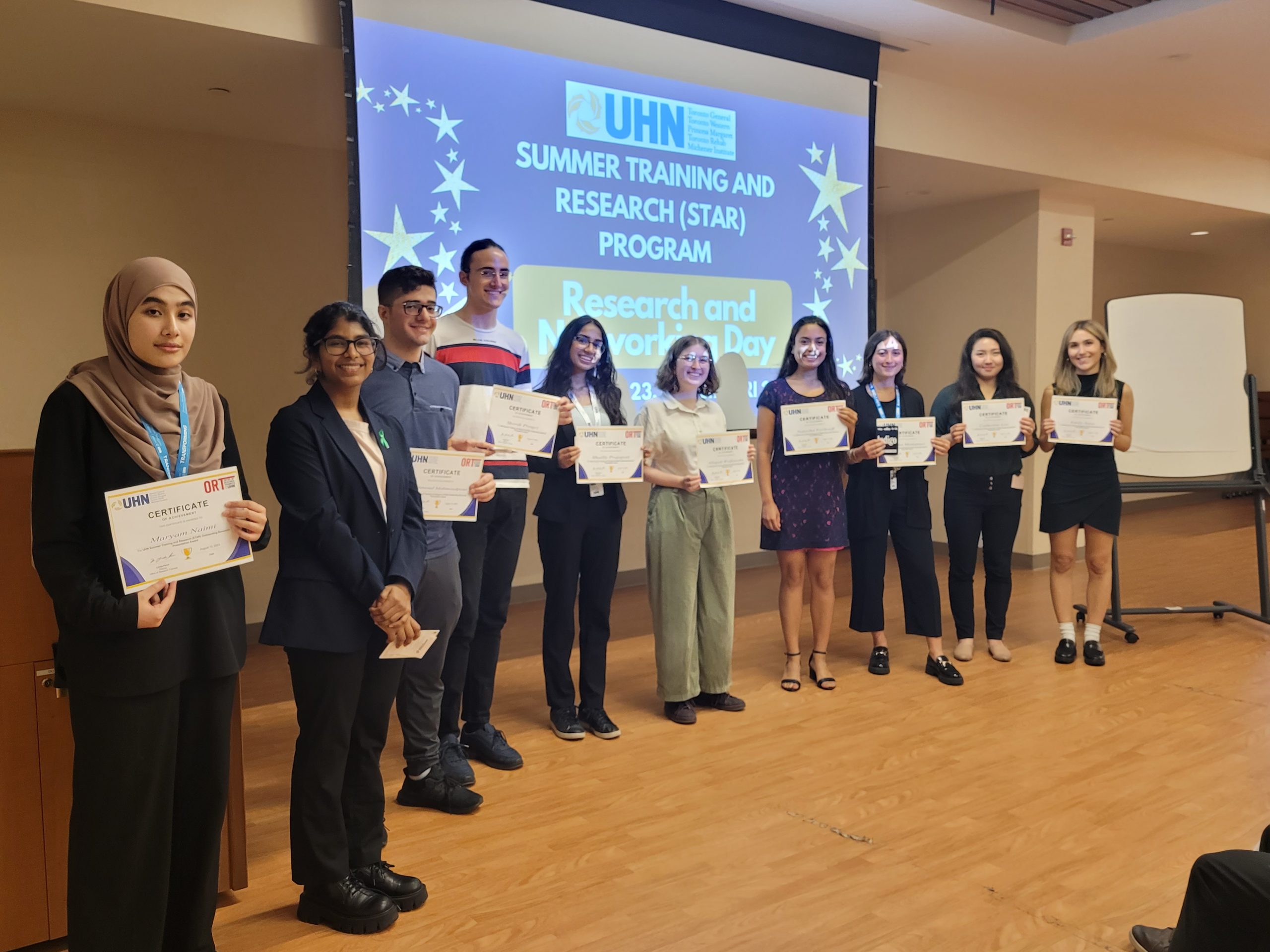 A group of nine individuals is posing for a photo at what appears to be a certificate award ceremony. They are standing in front of a presentation screen that reads "UHN SUMMER TRAINING AND RESEARCH (STAR) PROGRAM Research." Each person is holding a certificate with the UHN logo, signifying some achievement or completion within the program.